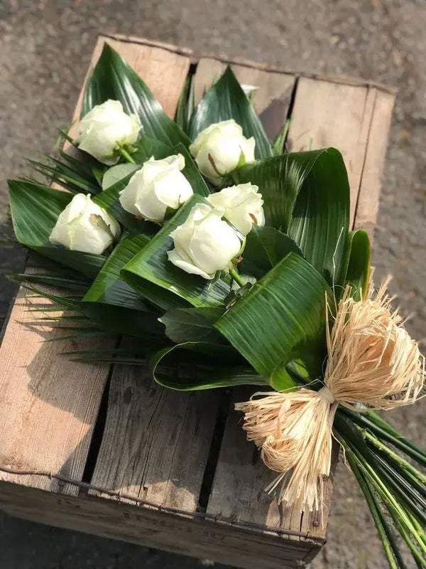 Rustic white rose funeral bouquet with tropical green leaves and raffia tie, ideal for expressing sympathy and honoring a loved one