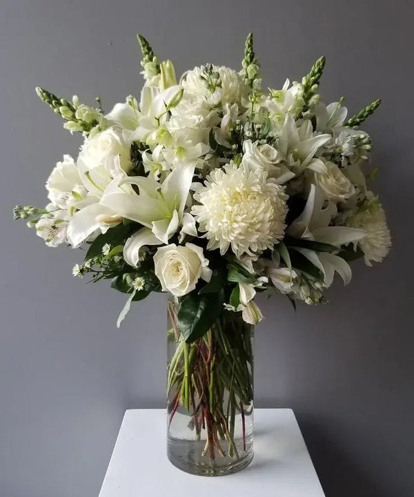 Elegant white funeral flower arrangement featuring roses, lilies, chrysanthemums, and snapdragons in a tall glass vase, symbolizing peace and remembrance.