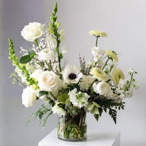 Delicate funeral bouquet with white roses, anemones, snapdragons, chrysanthemums, and ranunculus in a clear glass vase, perfect for honoring and remembering loved ones.