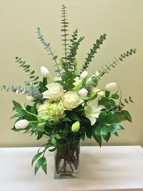 Elegant white funeral bouquet with roses, lilies, tulips, and eucalyptus in a glass vase – sympathy floral arrangement for funerals