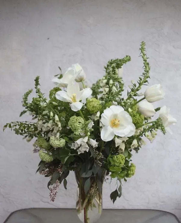 Elegant white funeral bouquet with tulips, lilies, and lush greenery in a glass vase – serene sympathy flower arrangement for memorial services.