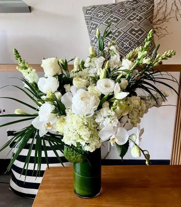 Elegant white funeral bouquet in a tall green vase featuring roses, hydrangeas, peonies, snapdragons, and orchids with lush greenery for memorial services.