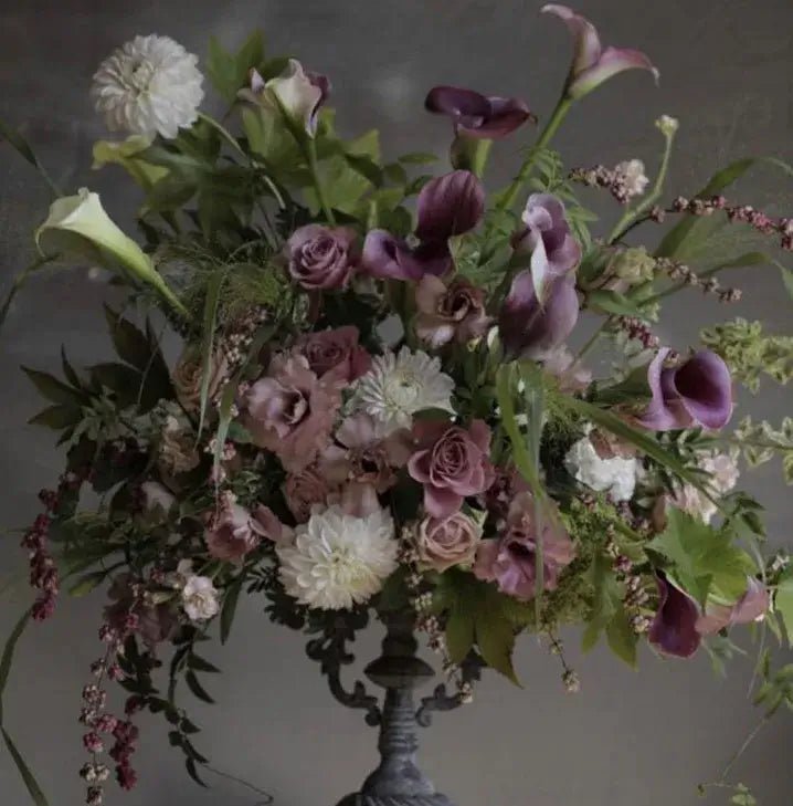 Elegant funeral bouquet with purple calla lilies, pink roses, white dahlias, and greenery in an ornate dark vase – sophisticated floral tribute for funerals and memorial services.