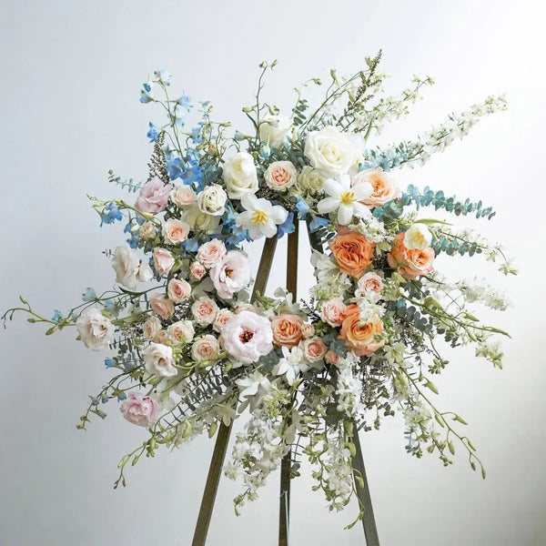 Elegant funeral wreath with pastel roses, blue delphiniums, white blossoms, and eucalyptus on a wooden stand – serene floral tribute for memorial services